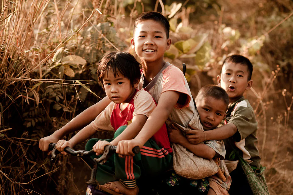 Four Thai boys sharing one bicycle through tall grass at golden hour